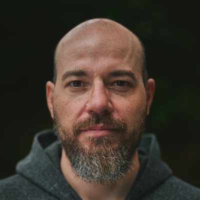 PJ Starling — a bald, bearded man in a grey hooded sweatshirt, looking straight at the camera against a dark background.